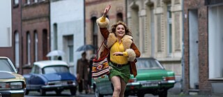 En scene fra filmen Köln 75, hvor en munter kvinde løber hen ad gaden med armen strakt opad.