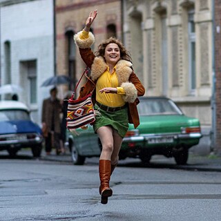 Eine Szene aus dem Film Köln 75 in der eine fröhliche Frau auf der Straße rennt und ihren Arm nach oben gestreckt hat