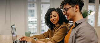 A smiling man and woman sitting in front of a laptop, looking at the man's phone