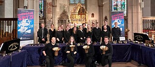 Die 20 Mitglieder des Handglockenchors Wiedensahl stehen in einer Kirche, vier von ihnen knieen im Vordergrund und halten Handglocken hoch, der Rest steht hinter einem Tisch, auf dem weitere Glocken zu sehen sind.