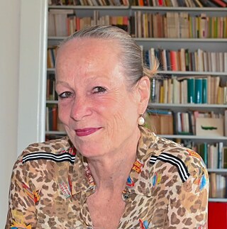 Πορτραίτο μιας μεσήλικης κυρίας. Έχει τα μαλλιά πιασμένα πίσω, φοράει σκουλαρίκια πέρλες και animal print πουκάμισο και χαμογελά ευγενικά. 