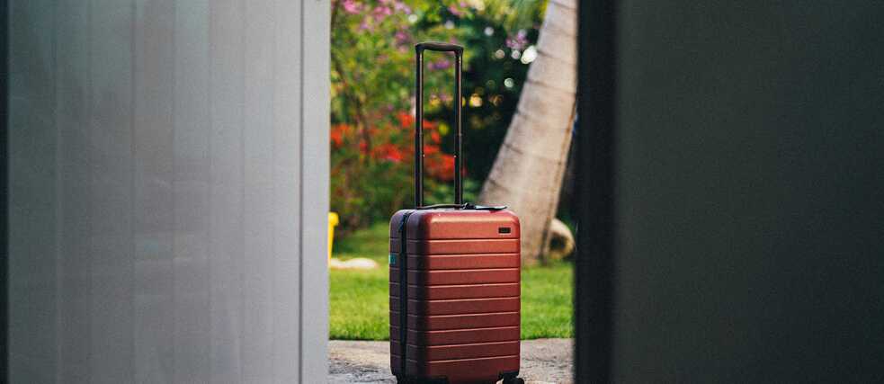 A red hardshell suitcase with wheels stands on a concrete surface just outside an open door, framed by a lush green garden with palm trees and vibrant flowers in the background.