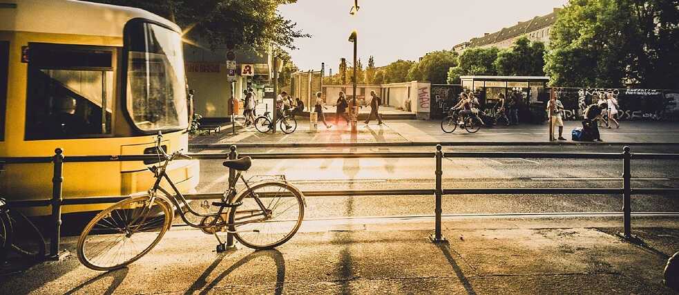 A city street at sunset with a tram, parked bicycles, and people walking or waiting.