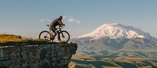 A cyclist stands on the edge of a rocky cliff, overlooking a vast green valley with rolling hills and a snow-capped mountain in the background under a clear blue sky © © Unsplash Cyclist on Cliff Edge with Snow-Capped Mountain View