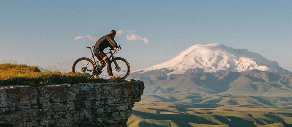 A cyclist stands on the edge of a rocky cliff, overlooking a vast green valley with rolling hills and a snow-capped mountain in the background under a clear blue sky