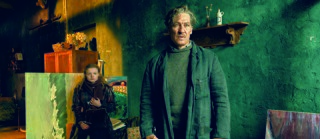 The image shows an older man and a woman in a dimly lit, artfully arranged room. The man stands in the foreground wearing dark clothing, while the woman stands behind him next to several paintings. The mood is serious, with soft lighting highlighting the scene. The green walls and background include art supplies and furniture.