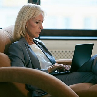 A woman dressed in a suit sits comfortably on a chair, engaged with her laptop.