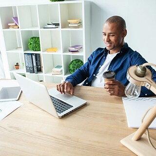 A man is seated at a desk, typing on a laptop, with a coffee nearby, immersed in his work.