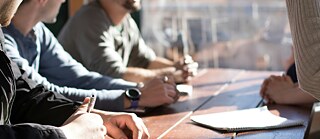 A group of people sitting at a table, holding notebooks and pens in their hands, engaged in conversation in a bright room. © © Unsplash Meeting at the Table with Notebook