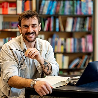 Ein Student lernt in der Bibliothek. Vor ihm liegen ein aufgeschlagenes Buch und ein Laptop. Er blickt lächelnd in die Kamera.