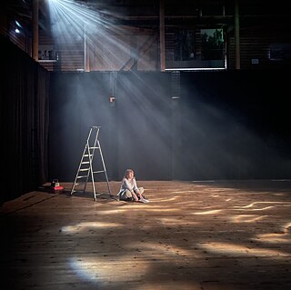 Eine Frau sitzt auf der Bühne und liest einen Text. Hinter ihr steht eine kleine Leiter.