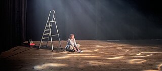 Eine Frau sitzt auf der Bühne und liest einen Text. Hinter ihr steht eine kleine Leiter.