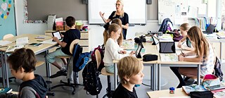 Eine Lehrerin erklärt Schülern im Klassenraum einen Sachverhalt. Im Hintergrund ein Smartboard.