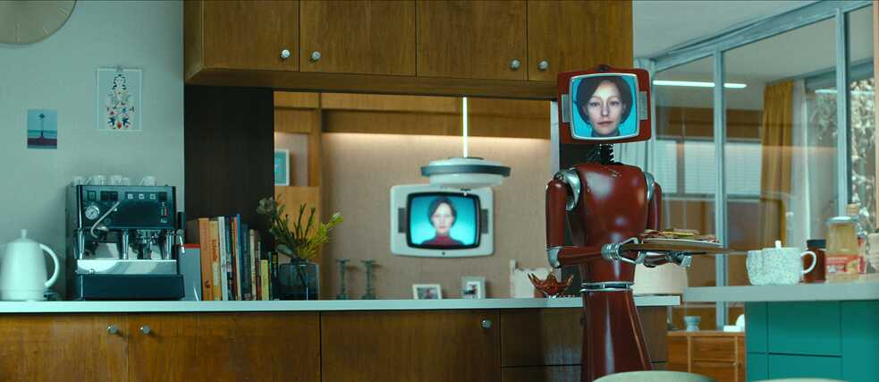 Vor dem Hintergrund einer 70er-Jahre Küchenzeile hält Cassandra - ein dunkelroter Roboter mit aufgesetzten Röhrenbildschirm, auf dem eine weiße Frau mit nackenlangen Haaren zu sehen ist - einen Teller mit belegten Brötchen. 
