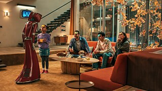 Against the backdrop of a 1970s living room, Cassandra - a dark red robot with an attached tube screen showing a white woman with neck-length hair - plays charades with the family.