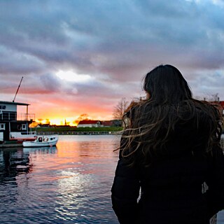 Eine junge Frau mit langen, dunklen Haaren steht am Hafenbecken und blickt auf das Wasser.
