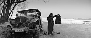 In a wintry landscape, a man next to a car is holding up a uniform 