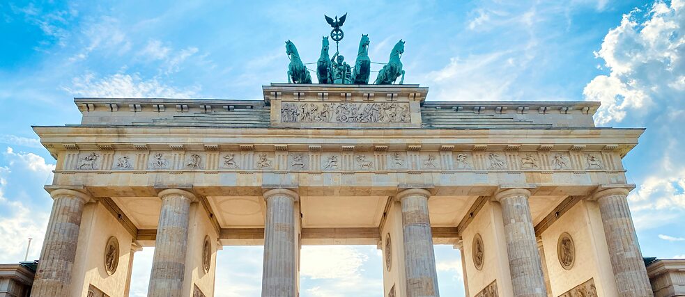h: Close-up of the Brandenburg Gate in Berlin, showing detailed carvings and the Quadriga sculpture against a blue sky.