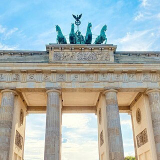 h: Close-up of the Brandenburg Gate in Berlin, showing detailed carvings and the Quadriga sculpture against a blue sky.