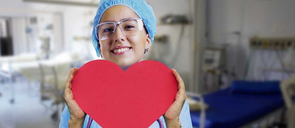 A healthcare professional in blue scrubs and a surgical cap holds a large red heart-shaped object in a hospital room with medical equipment and a bed in the background.
