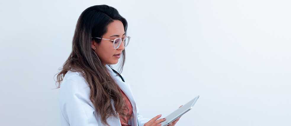 A doctor in a white coat reviews information on a tablet against a plain background.