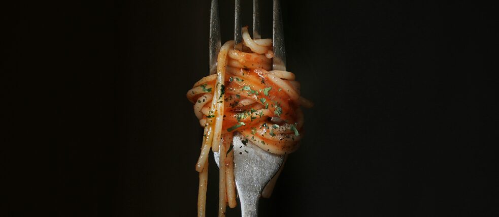 A close-up of a fork twirled with spaghetti in tomato sauce, garnished with herbs, against a black background.
