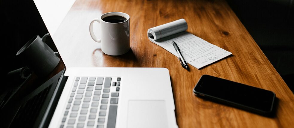 A wooden desk with a laptop, a cup of coffee, a notepad with handwritten notes, a black pen, and a smartphone. The workspace is well-lit, creating a cozy and focused atmosphere.