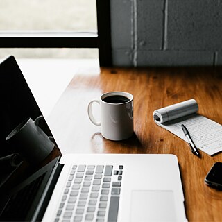A wooden desk with a laptop, a cup of coffee, a notepad with handwritten notes, a black pen, and a smartphone. The workspace is well-lit, creating a cozy and focused atmosphere.