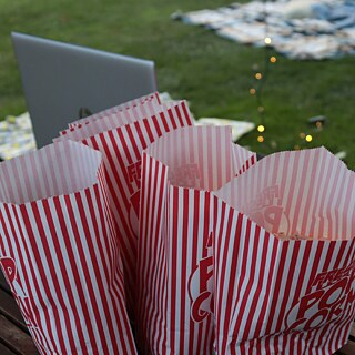 Mehrere rot-weiß gestreifte Popcorn-Tüten auf einem Holztisch, während eine Hand danach greift. Im Hintergrund sind grünes Gras, Lichterketten und eine gemütliche Outdoor-Atmosphäre zu sehen, was auf einen Filmabend oder eine gesellige Veranstaltung hindeutet.