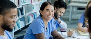 In the center is a smiling young nurse with dark hair, wearing a blue coat and a brown undershirt. Her colleague can also be seen. He is looking at notes on the table. They are surrounded by a shelf of books and files in the background.