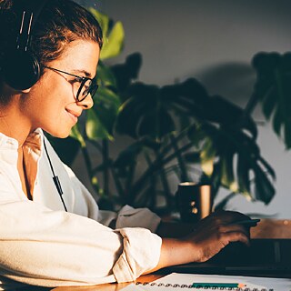 A person wearing headphones sits at a desk with a laptop, a mug, and a notebook. Green plants are in the background, and warm light fills the room, creating a cozy work atmosphere.
