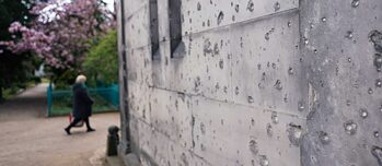 Bullet holes on a building in the Dorotheenstädtischer Friedhof cemetery in the foreground, a person and a blossoming tree in the background.