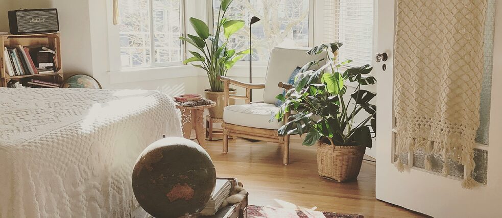 A cozy bedroom featuring a white bed with a textured blanket, a globe on a vintage suitcase, a red patterned rug, large windows with wooden blinds, several potted plants, a hanging macrame plant holder, a bookshelf with books, and a woven wall hanging near an open door.