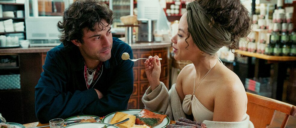 Vor der unscharfen Kulisse einer Ladentheke und von Regalen mit Lebensmitteln reicht Saba (Saffron Coomber) Samuel (Aaron Altaras) eine Gabel mit einer Bullette, die an Königsberger Klopse erinnert – während sich die beiden dabei verliebt in die Augen sehen.