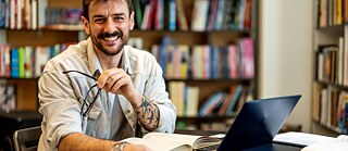 Ein Student lernt in der Bibliothek. Vor ihm liegen ein aufgeschlagenes Buch und ein Laptop. Er blickt lächelnd in die Kamera.