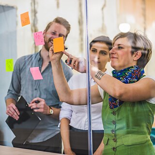 Zwei Frauen und ein Mann stehen vor einer Glaswand, an der Post-its aufgehängt sind, die Frau links im Bild schreibt gerade etwas auf einen Zettel, der Mann links im Bild lacht. 