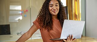 Eine junge Frau steht im Büro, sie trägt ein Laptop auf dem Arm und lächelt in die Kamera.