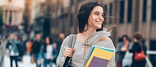 Eine junge Frau auf dem Campus. Sie hält Bücher im Arm und lächelt glücklich.
