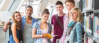 Junge Studenten stehen in einer Bibliothek und blicken lächelnd in die Kamera.
