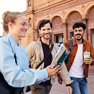 Drei junge Erwachsene, eine Frau und zwei Männer, stehen fröhlich vor einem rötlichen historischen Gebäude. Die Frau im Vordergrund hält eine Broschüre in der Hand und zeigt ihren beiden männlichen Begleitern etwas darauf. Alle drei wirken entspannt, neugierig und gut gelaunt.