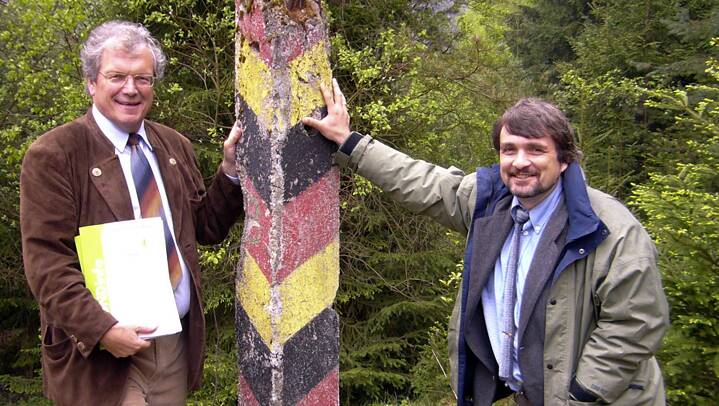 Links und rechts von einem Grenzpfahl in Deutschlandfarben stehen zwei Männer. Im Hintergrund ist Wald zu sehen.