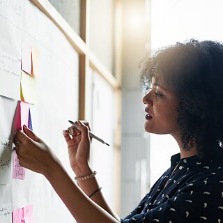 Eine junge Frau steht an einer Pinnwand mit bunten Post-its und macht weitere Notizen.
