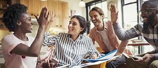 Lockere Arbeitsatmosphäre. Junge Studierende lernen gemeinsam mit klassischen und digitalen Arbeitsunterlagen. Sie geben sich „High fives“. Sie haben Spaß.