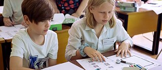 In einem Klassenraum sitzen ein Junge und ein Mädchen gemeinsam an einem Tisch. Beide sind tief in ihre Aufgaben vertieft und wirken sehr konzentriert.