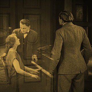 Sepia-coloured silent film scene in which a man in back view sees a woman sitting at a piano and a man bent over her in ghostly transparency
