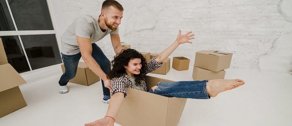  A man using a box like a shopping cart to push his happy wife across the floor of their new apartment.