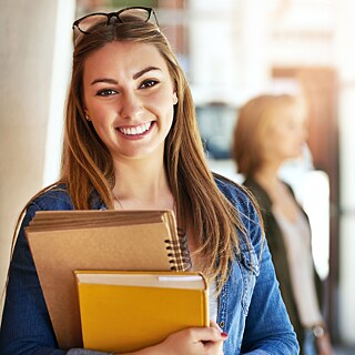 Eine Frau hält ein Buch und einen Ringordner in der Hand und lächelt in die Kamera. Im Hintergrund unterhalten sich Studentinnen.
