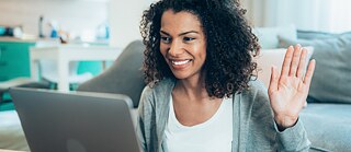 	Eine junge Frau sitzt zu Hause vor dem Sofa und arbeitet lächelnd am Laptop. Sie lächelt und winkt in Richtung Bildschirm.