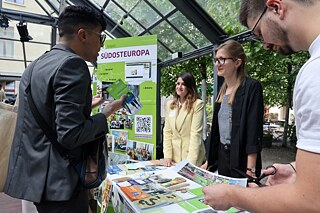 A man stands at the table of the Southeast Europe region and asks two employees of the Goethe-Institut for information