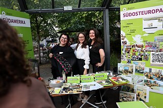Three women stand behind a table with information material and laugh